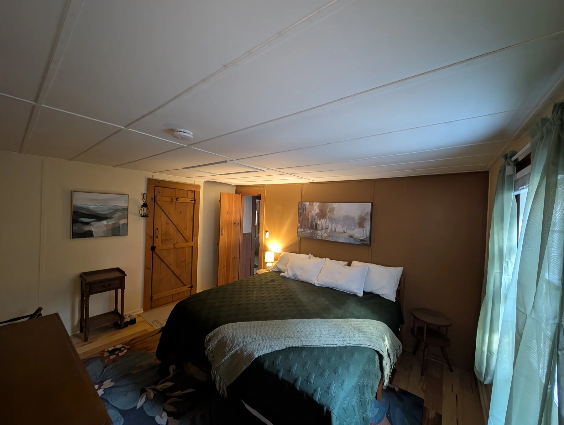 Bedroom with a large bed, layered green bedding, bedside tables, and a forest-facing window